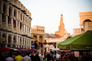A large crowd of people walking through the souq waqif streets, with restaurants from left and right. There is the finar mosque in the background.