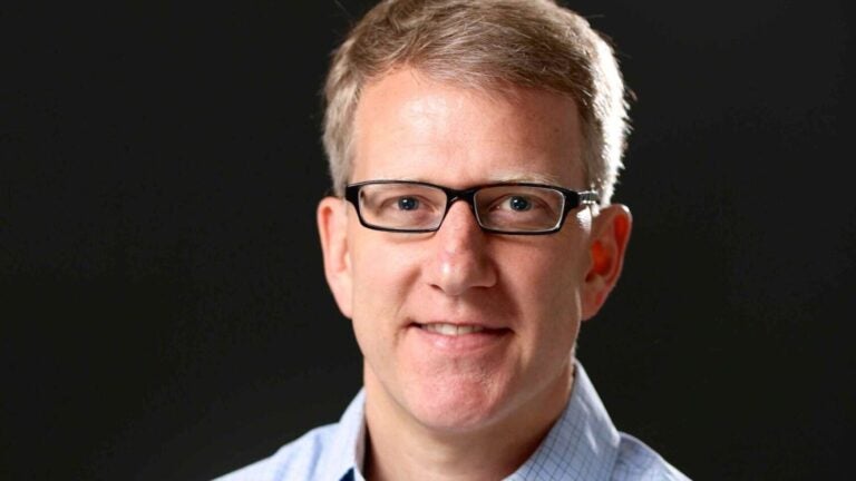 Image of the speaker Sebastian Abbot in a light blue shirt, with his arms crossed and wearing black, rectangular glasses.