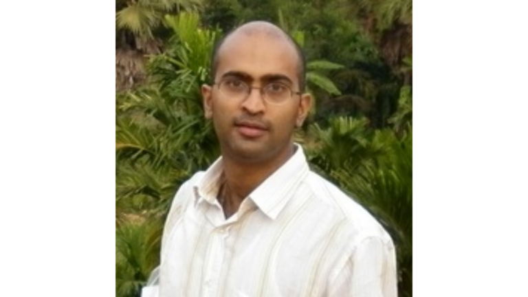 Picture of Jason in a white collared shirt, wearing glasses and looking at the camera, in front of a green plant wall