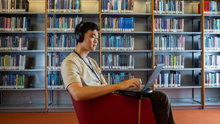 Man sitting with laptop in library with bookshelf in the background.