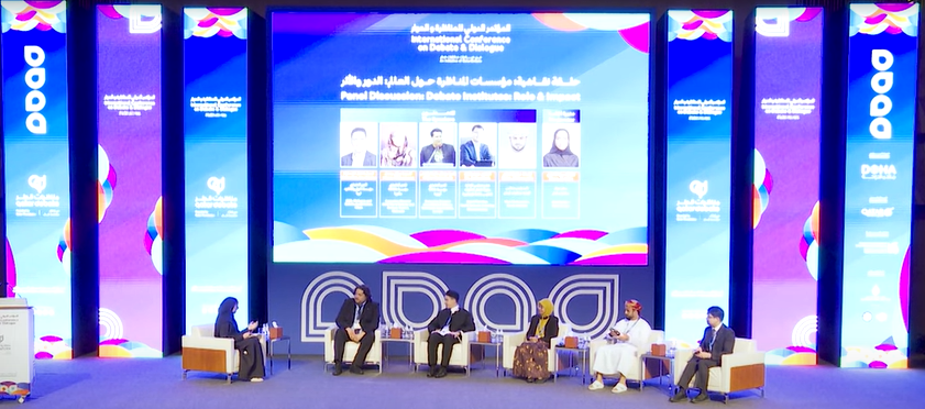 Stage at International Conference on Debate and Dialogue showing five panelists with Moza Al Hajri seated to the right leading the conversation. Conference branding screens are behind the panelists showing blue and purple conference branding.