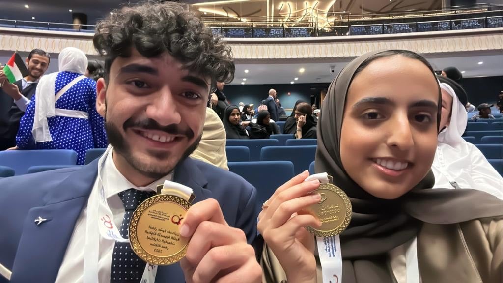 Hamza AlSioufy and Moza AlHajri seated side by side holding up gold medals from their win the 2nd Arabic Debate Championship, Oman