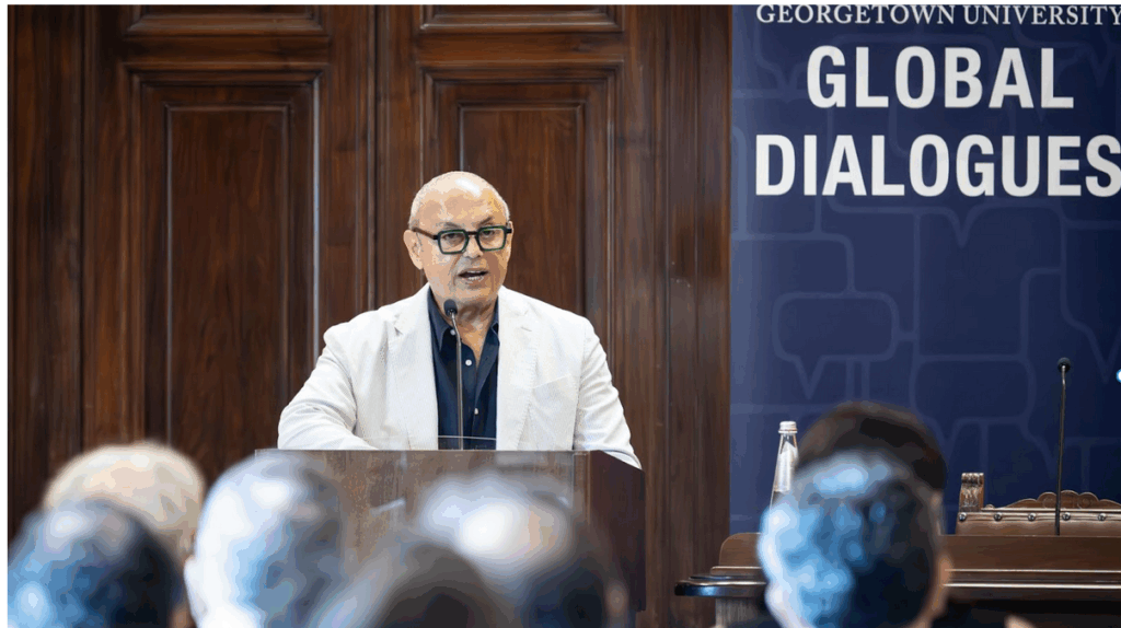 Dean Masri stands at podium with Global Dialogues banner behind addressing a sea of heads. He is wearing a white blazer, black shirt and glasses.