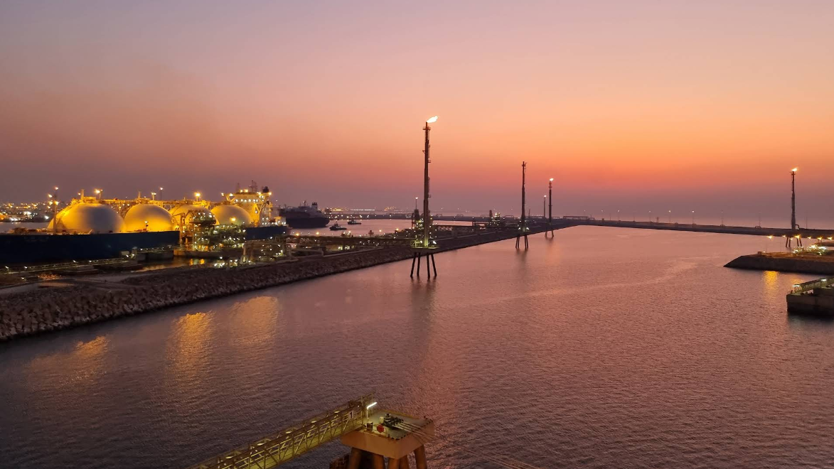 Ras Laffan Port at sunset with lit up LNG ship and gas egress towers topped with fire and water reflecting orange and purple afterglow of sunset.
