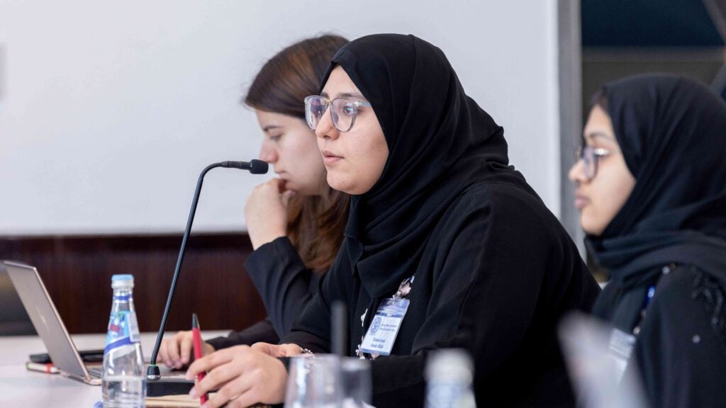 Girl in black hijab and glasses speaks into a microphone in profile view.