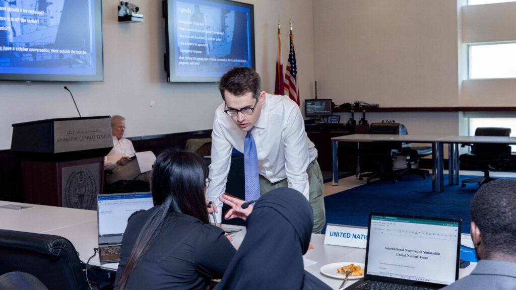 Man in a tie, a diplomat, leans over a table of hijabi girls in a classroom setting with white tables and screens on the wall. Computer screen on the table in front of the girls shows that it is a crisis negotiation exercise.